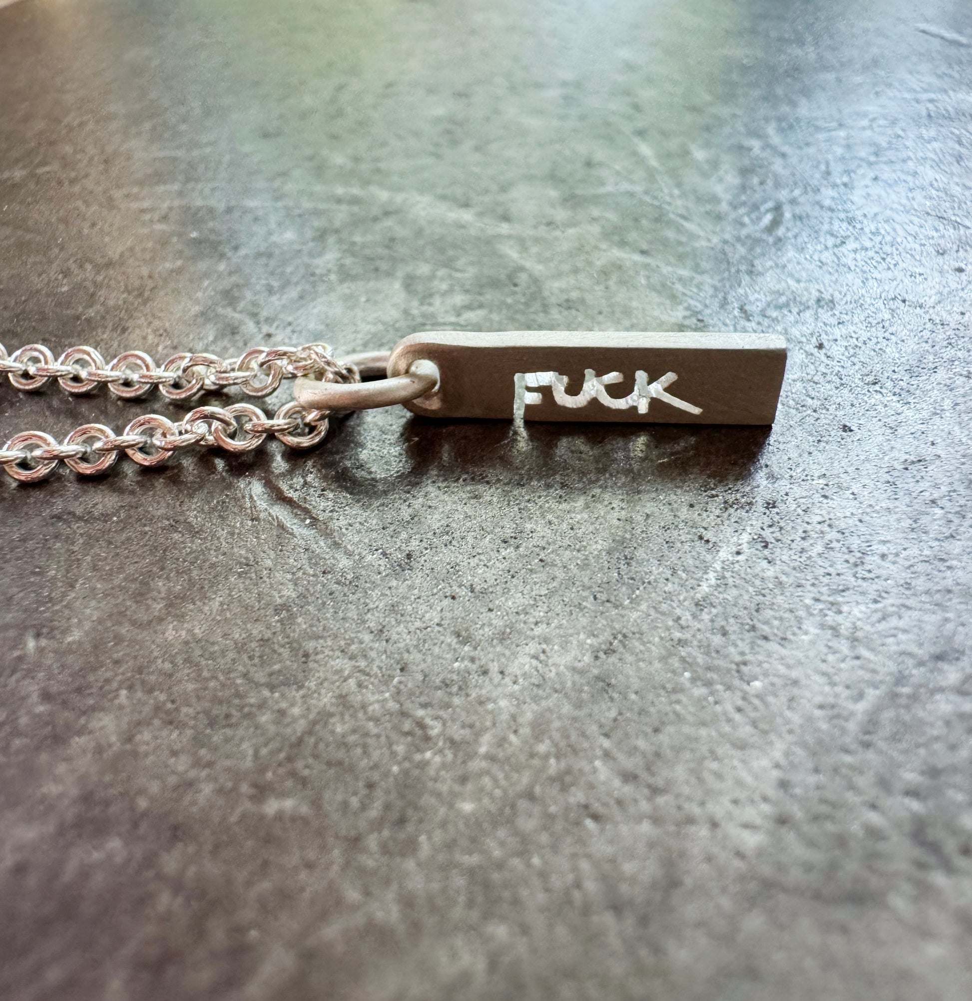 Silver tag style charm with "FUCK" cut into it, shown on the surface of my anvil.