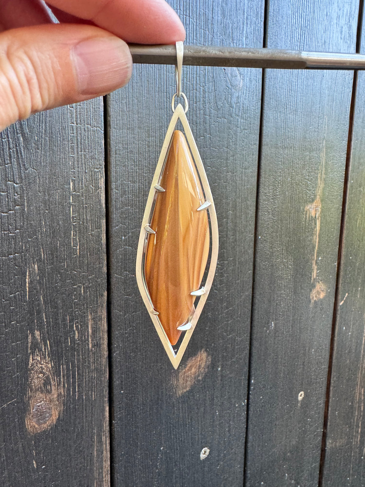 Pendant with caramel and tan colored stone with elongated tapered shape set in a frame with square wire and sculpted prongs. Held against the charred wood siding of my studio. The stone is an Oregon fire opal.